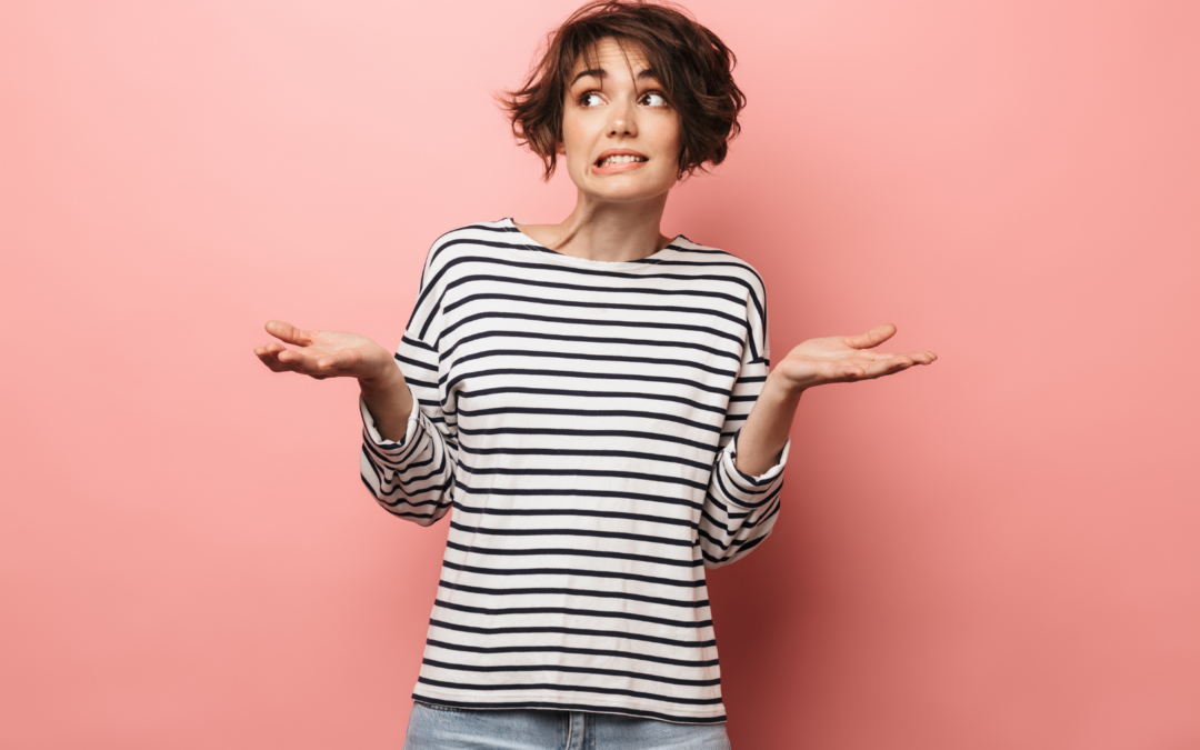 Young woman in black and white striped shirt stands in front of a pink wall with a confused expression on her face what lifting her hands in a questioning pose.
