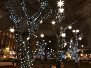 Trees in a plaza decorated with blue & white twinkle lights and large white stars.