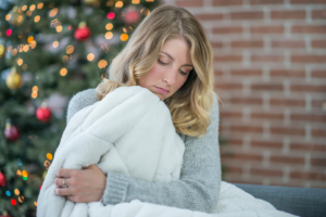 Woman sits in front of a Christmas tree, sadly clutching a blanket.