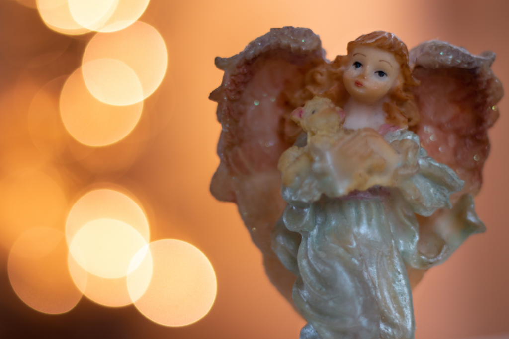 Ceramic Christmas Angel stands a long with blurred warm lights behind it.