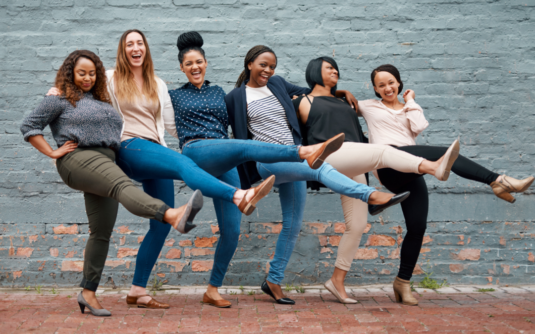 Six multi-ethnic professional women join in a kick-line, laughing and smiling.