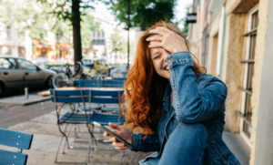 Red-haired woman sits at a bistro table on a city sidewalk. She hold her phone and laughs with her hand on her forehead.
