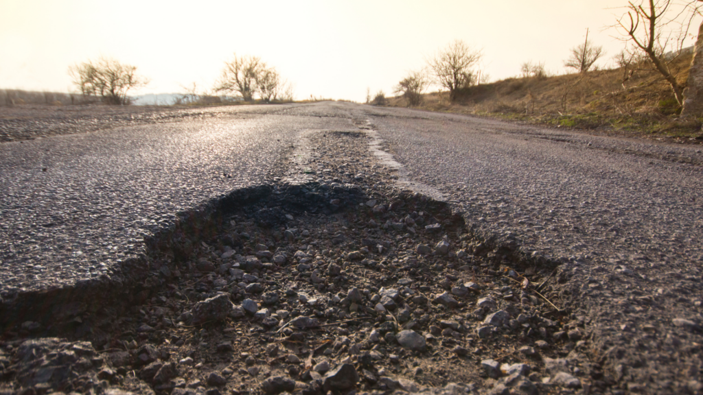 The sun shines on a patched-together road that stretches to the horizon. In the foreground is a large pothole.