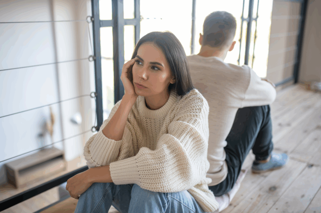 Woman in a cream sweater sitting on the floor, looking upset and distant while sitting back-to-back with a man, symbolizing marital tension or emotional disconnect.