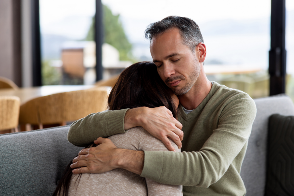 Man gently embracing a grieving woman on a sofa, eyes closed, offering comfort and support after loss in ministry.
